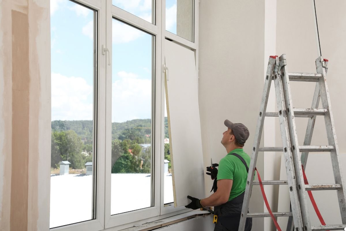 A man on a ladder painting a window, focused on his task with paintbrush in hand and a paint can nearby.